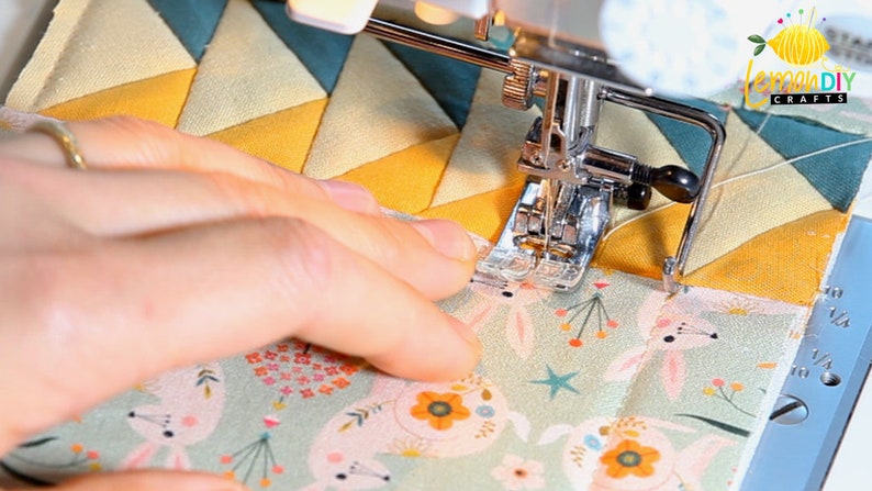 May include: A close-up of a sewing machine needle stitching two pieces of fabric together. The top fabric is a yellow and blue geometric pattern, and the bottom fabric is a green fabric with a white bunny rabbit pattern. The sewing machine foot is a walking foot.