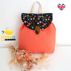 Puede incluir: Una mochila de lona de color coral con una solapa de estampado floral y un cierre de hebilla de latón. La mochila tiene un cordón de ajuste blanco y correas ajustables.