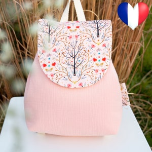 Peut inclure: Un sac à dos rose avec un rabat orné d'un motif floral avec des arbres, des cœurs et des papillons. Le rabat est fermé par une sangle blanche.