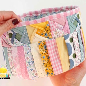 Könnte beinhalten: Ein handgefertigter Stoffkorb mit Patchwork-Design. Der Korb zeigt verschiedene gemusterte Stoffe in Rosa, Gelb, Blau und Grün. Der Rand ist mit rosa und weiß gestreiftem Stoff gefüttert. Der Korb wird von einer Person gehalten.