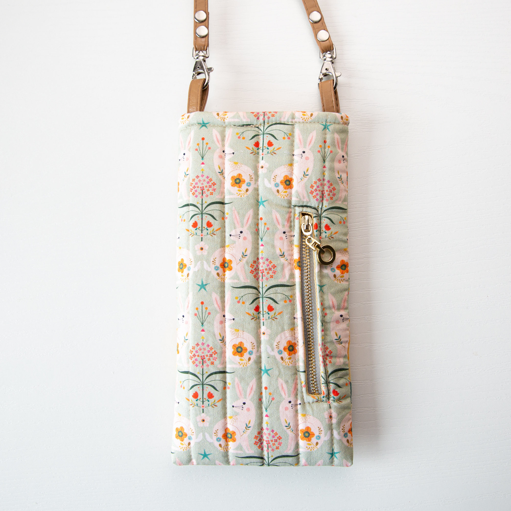 May include: A light green quilted crossbody bag with a zippered pocket and a brown strap. The bag features a repeating pattern of white bunnies and colorful flowers.