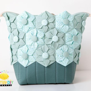 Könnte beinhalten: Eine türkisfarbene Reißverschlusstasche mit gestepptem Boden und einer Vorderseite aus Origami-Blumen aus Stoff. Die Blumen sind hellblau mit weißen Punkten und haben einen hellblauen Knopf in der Mitte.