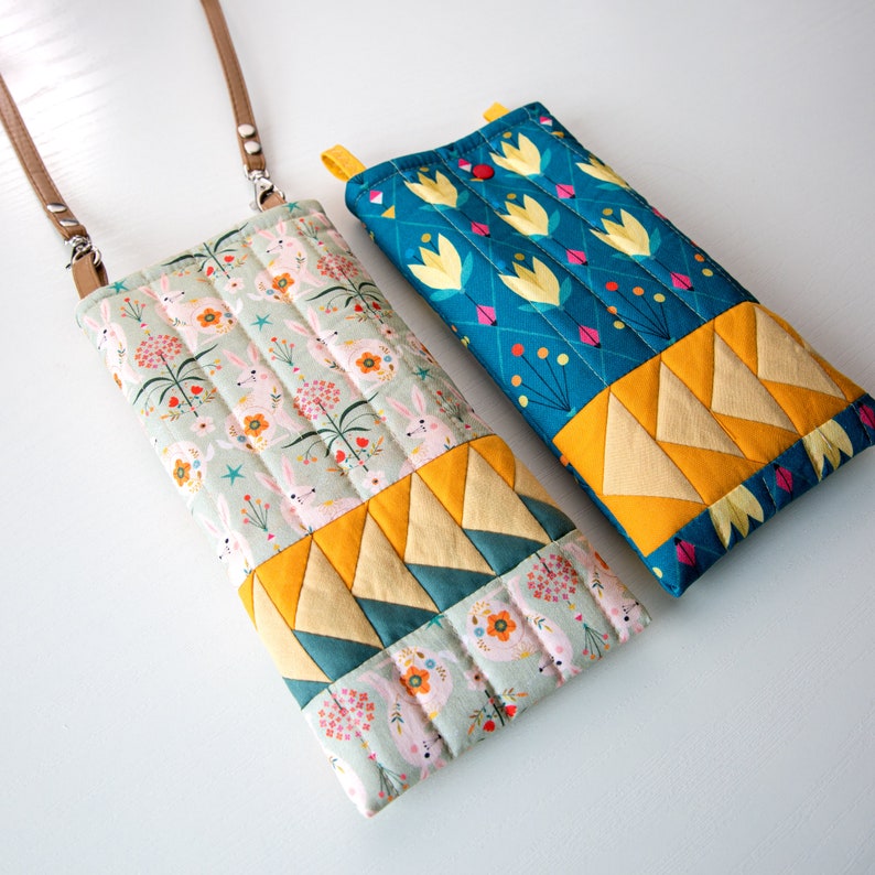 May include: Two quilted phone cases, one with a floral pattern in shades of blue and yellow, and the other with a pattern of bunnies and flowers in shades of green, pink, and yellow.