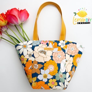 Könnte beinhalten: Eine Tragetasche mit Blumenmuster mit marineblauem Hintergrund und großen orangefarbenen, weißen und gelben Blumen. Die Tasche hat senfgelbe Griffe und ein kleines Lederetikett. Ein Strauß roter und gelber Tulpen befindet sich im Inneren.