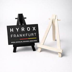 Könnte beinhalten: Zwei kleine Holzstaffeleien, eine schwarze und eine aus Naturholz. Die schwarze Staffelei trägt ein schwarzes Schild mit weißem Text: "HYROX FRANKFURT" und "OPEN_DOUBLES_SEASON_2025/2026". Die andere Staffelei ist aus hellem Holz.