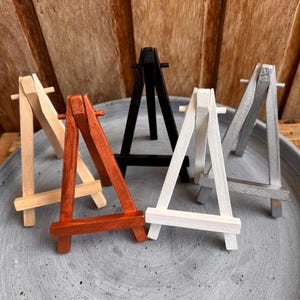 Könnte beinhalten: Eine Sammlung kleiner Holzstaffeleien in verschiedenen Farben, darunter Naturholz, Orange, Schwarz, Weiß und Silber. Die Staffeleien sind auf einer grauen, kreisförmigen Oberfläche angeordnet, ideal für die Präsentation kleiner Kunstwerke oder Fotos.