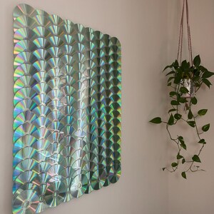 Peut inclure: Une œuvre d'art murale composée de CD irisés disposés en éventail. Les CD reflètent un spectre de couleurs, créant un effet chatoyant. L'œuvre est fixée au mur, avec une plante suspendue en arrière-plan.