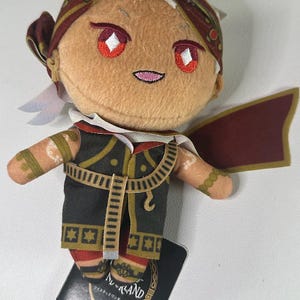 May include: A plush doll with a tan face, red eyes, and a small smile. It wears a dark outfit with gold accents, a red and gold headpiece, and a matching cape. A black tag with Japanese text is attached.