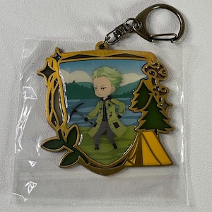 May include: A gold-colored metal keychain featuring a cartoon character in a green coat, holding a pickaxe. The character is set against a landscape with a lake and mountains. The keychain has a clasp for easy attachment.