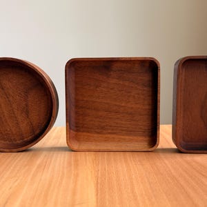 May include: Three wooden serving trays in different shapes: a round tray, a square tray, and a rectangular tray. The trays are made of a dark brown wood and sit on a light brown wooden surface.