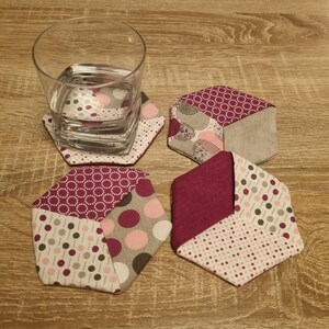Könnte beinhalten: Set aus vier sechseckigen Stoffuntersetzern. Jeder Untersetzer hat ein Patchwork-Design in Burgunderrot, Rosa, Grau und Weiß. Ein klares Glas mit Wasser steht auf einem der Untersetzer.