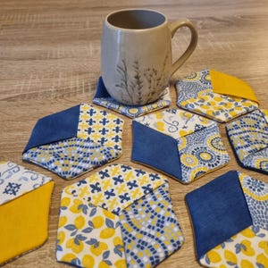Könnte beinhalten: Eine beige Tasse steht auf einem gemusterten Untersetzer, umgeben von mehreren sechseckigen Untersetzern. Die Untersetzer bestehen aus blauen, gelben und weißen Stoffen mit verschiedenen Mustern, darunter Zitronen und Blumen. Sie schützen Oberflächen vor Getränken.