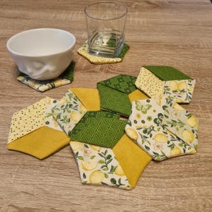 Könnte beinhalten: Eine Sammlung handgefertigter sechseckiger Stoffuntersetzer in verschiedenen Mustern. Die Untersetzer haben gelbe, grüne und weiße Stoffe mit Zitronen-, Blumen- und geometrischen Designs. Eine weiße Schüssel und ein klares Glas stehen auf passenden Untersetzern.