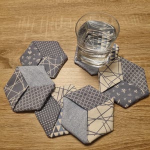 Könnte beinhalten: Sechs sechseckige Stoffuntersetzer in verschiedenen Grau- und Off-White-Mustern. Ein Glas Wasser steht auf einem Untersetzer. Die Muster umfassen Punkte, geometrische Linien und nautische Symbole. Auf einer Holzoberfläche angeordnet.