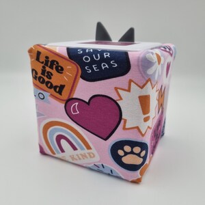 Op de afbeelding: Een roze stoffen kubus met verschillende kleurrijke pleisters en tekst, waaronder "Life is Good", "Save Our Seas" en "Be Kind".