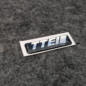 May include: A rectangular black and silver emblem with the letters "TTEI" in white. The emblem is on a white background and is set against a gray, textured surface.