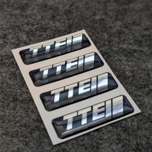 May include: Four glossy black and silver TTEII stickers arranged on a white backing. The stickers have a raised, three-dimensional appearance, with the letters TTEII in a bright silver color.