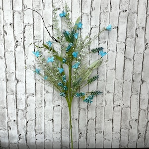 May include: A decorative floral stem featuring light blue flowers, small teal berries, and green foliage. The artificial arrangement includes a dark, thin branch and is set against a white brick wall.