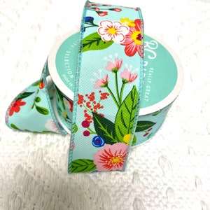 May include: A roll of light blue floral ribbon with a repeating pattern of red, pink, and white flowers, green leaves, and blue berries. The ribbon is on a white surface.