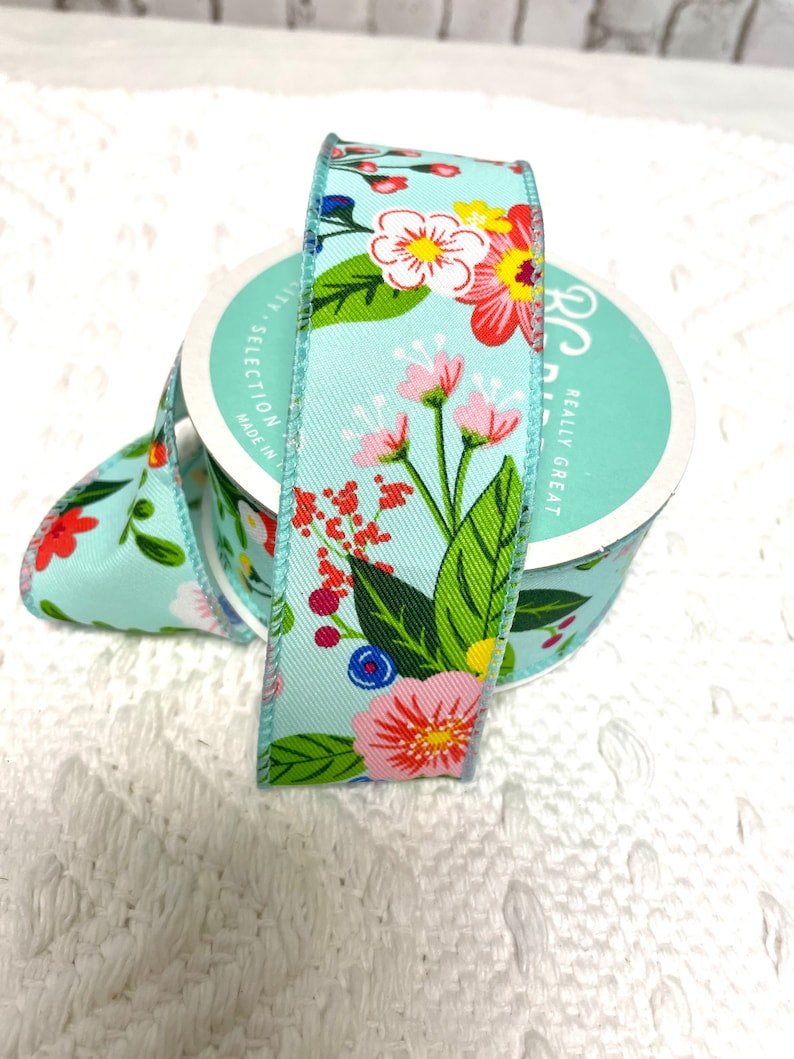 May include: A roll of light blue floral ribbon with a repeating pattern of red, pink, and white flowers, green leaves, and blue berries. The ribbon is on a white surface.