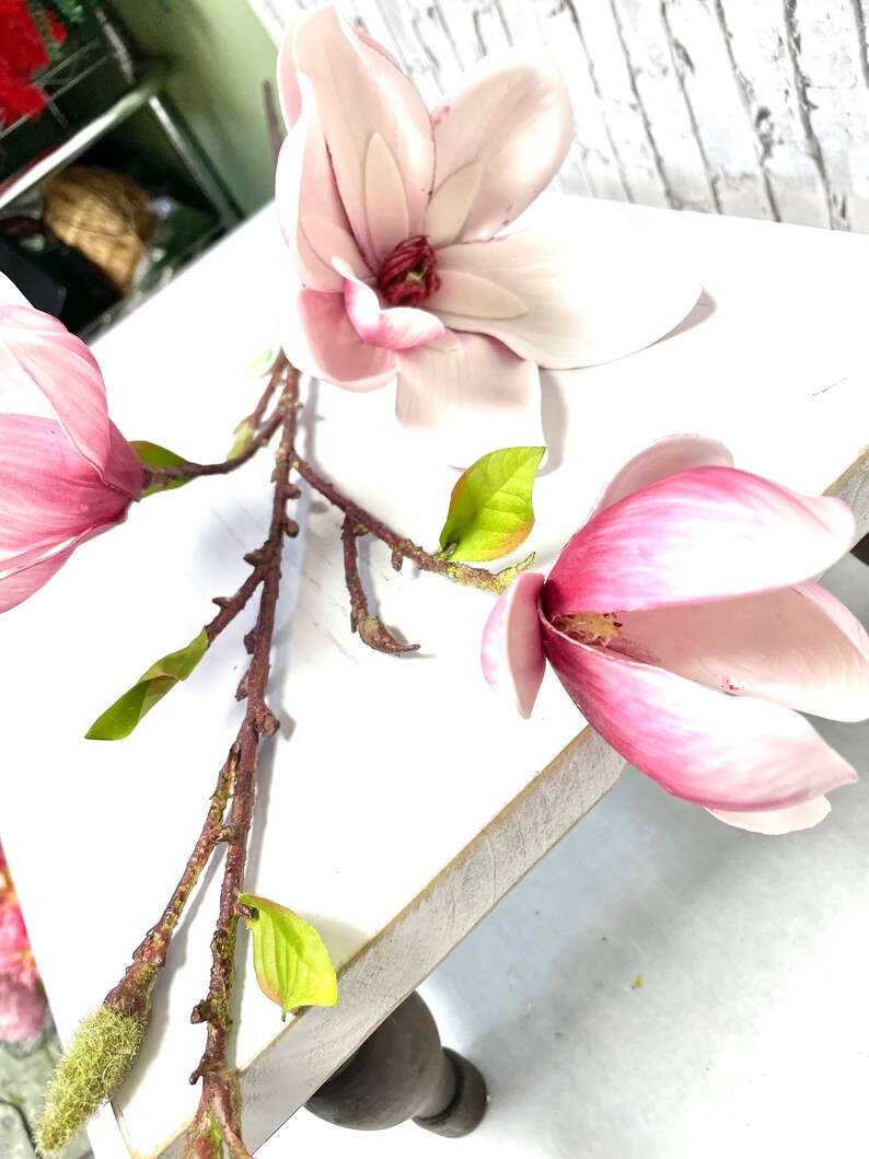May include: A pink magnolia flower branch with three blossoms and green leaves. The branch is on a white wooden surface.