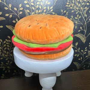 May include: A plush burger toy with orange, green, red, and brown layers, resembling a hamburger. The toy is displayed on a small, white, three-legged stand. The background features a black and gold floral pattern.