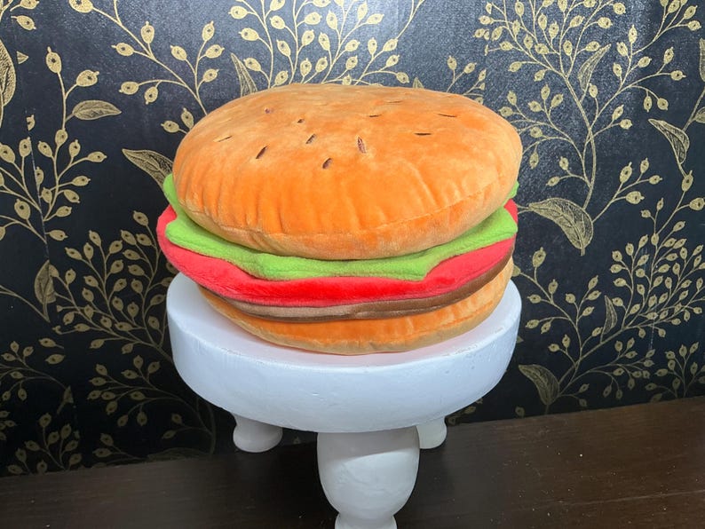 May include: A plush burger toy with orange, green, red, and brown layers, resembling a hamburger. The toy is displayed on a small, white, three-legged stand. The background features a black and gold floral pattern.