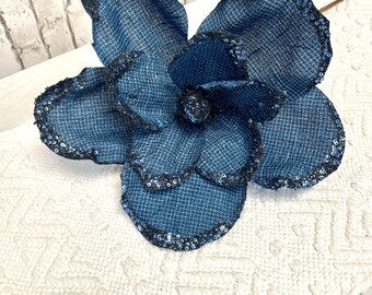 May include: A large blue fabric flower with a sequined edge. The flower is made of a textured material and has a ruffled appearance.