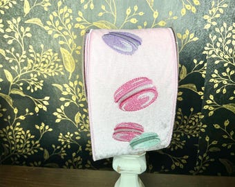 Puede incluir: Una toalla de mano rosa con macarons bordados en morado, rosa y verde. La toalla se exhibe sobre un soporte blanco adornado, sobre un fondo oscuro con un estampado floral dorado. El diseño evoca un tema de panadería o postre.