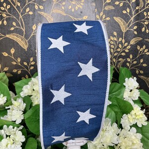 May include: A navy blue ribbon with white stars and a white trim. The ribbon is displayed in front of white flowers and green leaves. The background is a dark floral pattern.