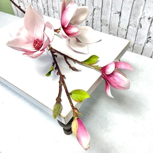 May include: A white wooden tray with a pink magnolia flower arrangement. The flowers are arranged in a natural, flowing style, with the stems extending beyond the edge of the tray. The tray is sitting on a white surface.