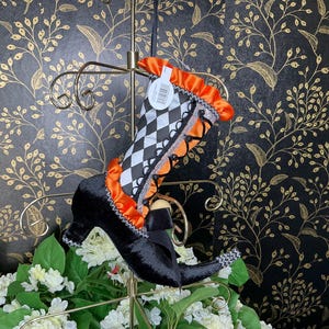 May include: A decorative Halloween witch boot with a black velvet shoe and a checkered pattern on the leg. The boot features orange and white accents, a black bow, and is displayed on a gold stand. The background has a black and gold floral design.