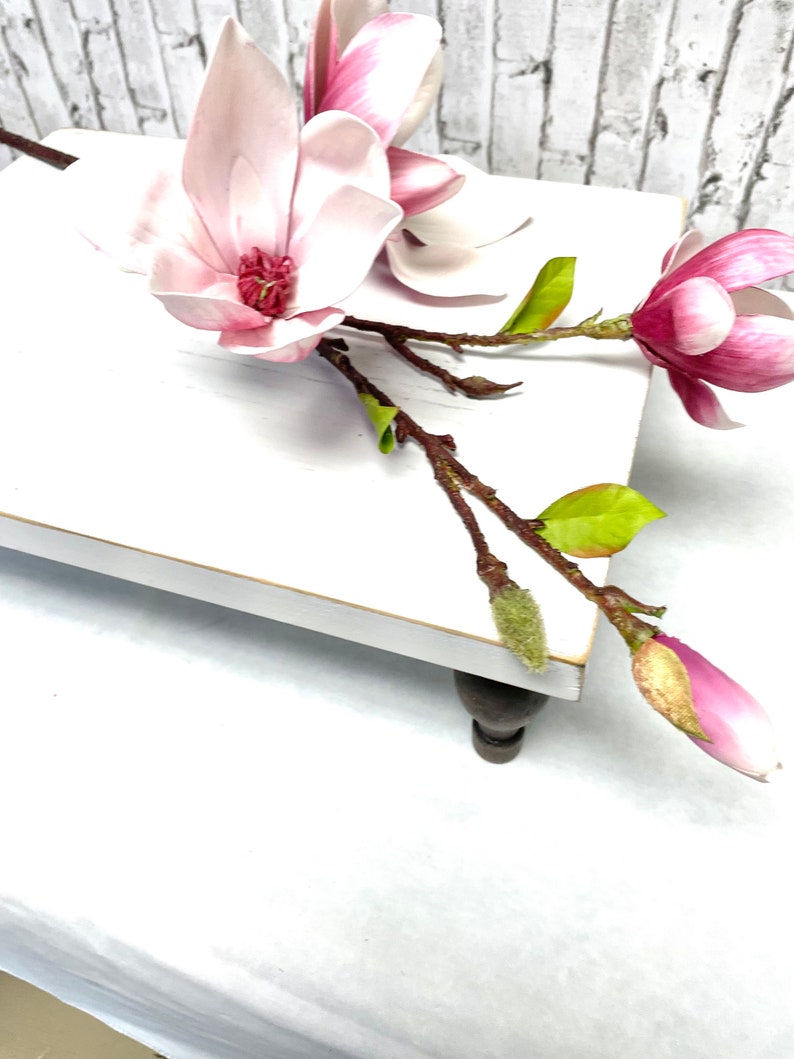 May include: A white wooden pedestal with a pink magnolia flower arrangement. The pedestal has turned wooden legs and a white painted surface.