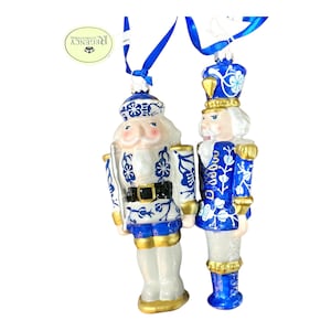 May include: Two decorative nutcracker ornaments. One is white with blue floral designs, a black belt, and gold accents. The other is blue with white floral designs and gold accents. Both have blue ribbons for hanging.