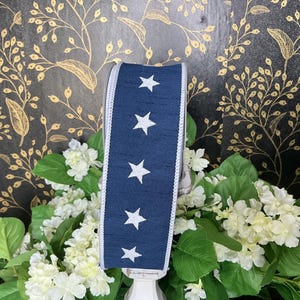 May include: A navy blue ribbon with white stars and a white border. The ribbon is displayed on a white stand, surrounded by white flowers and green leaves, against a black and gold floral background.