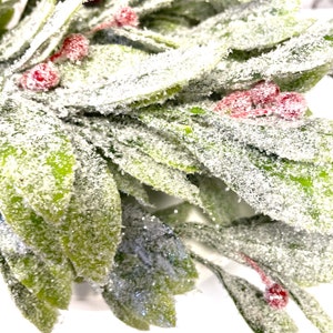 Puede incluir: Primer plano de un arreglo festivo con hojas verdes espolvoreadas con purpurina blanca y peque&ntilde;as bayas rojas. Las hojas tienen un aspecto esmerilado, creando una est&eacute;tica invernal. El arreglo es probablemente para la decoraci&oacute;n de temporada.