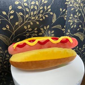 May include: A plush hot dog toy with a pink sausage, red relish, and yellow mustard, on a yellow bun. A fun, food-themed item.