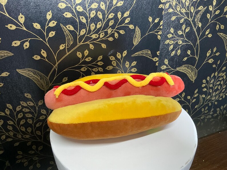 May include: A plush hot dog toy with a pink sausage, red relish, and yellow mustard, on a yellow bun. A fun, food-themed item.