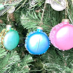 May include: Three Christmas ornaments in shades of teal, blue, and pink hang on a green Christmas tree. The teal ornament has gold glitter, the blue has blue beads, and the pink has white lace.