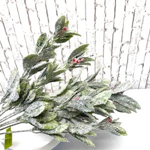 Puede incluir: Un ramo de vegetaci&oacute;n artificial con hojas verdes y peque&ntilde;as bayas rojas, espolvoreado con purpurina blanca. Los tallos est&aacute;n dispuestos en un cuenco blanco con pedestal, contra una pared de ladrillos blancos.