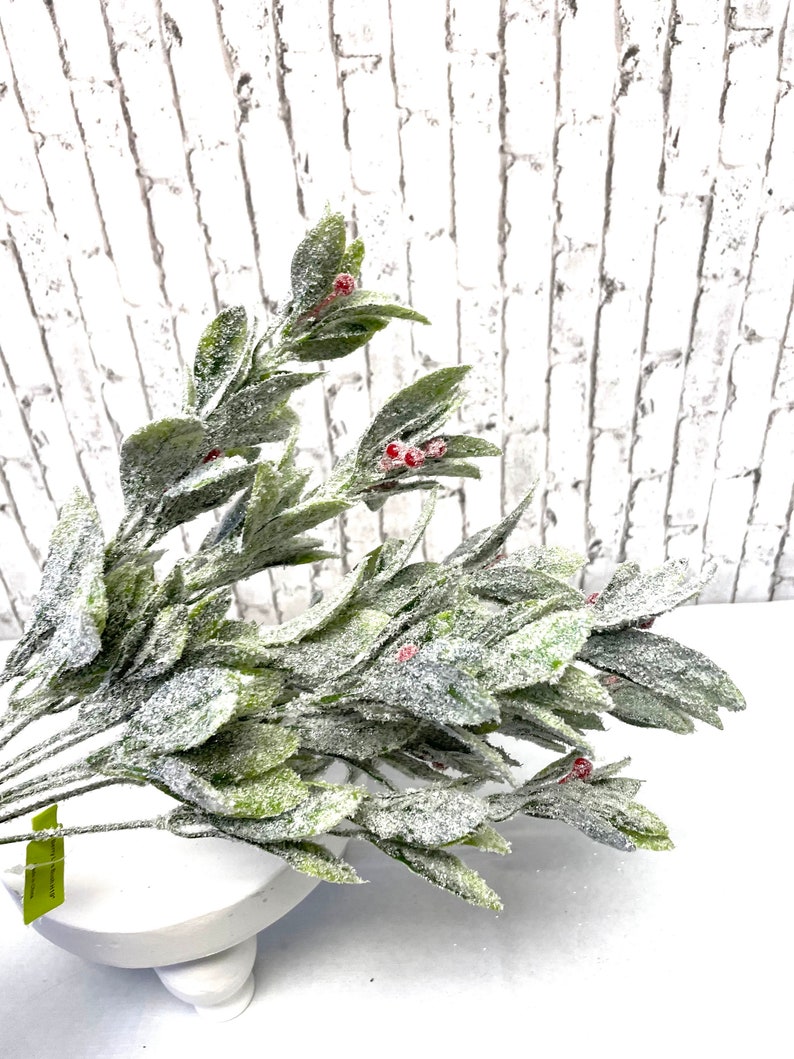 Puede incluir: Un ramo de vegetaci&oacute;n artificial con hojas verdes y peque&ntilde;as bayas rojas, espolvoreado con purpurina blanca. Los tallos est&aacute;n dispuestos en un cuenco blanco con pedestal, contra una pared de ladrillos blancos.