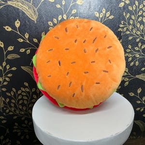 May include: A plush burger pillow with an orange top bun featuring brown seed details. The burger has a red patty, green lettuce, and is displayed on a white pedestal. The background is a black wall with a gold floral pattern.