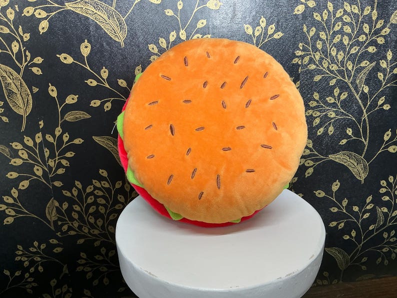 May include: A plush burger pillow with an orange top bun featuring brown seed details. The burger has a red patty, green lettuce, and is displayed on a white pedestal. The background is a black wall with a gold floral pattern.
