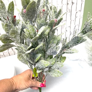 Puede incluir: Un ramo de vegetaci&oacute;n artificial con hojas verdes y peque&ntilde;as bayas rojas, cubierto con un material blanco similar a la nieve. Los tallos son verdes y las hojas son ovaladas. Fondo de pared de ladrillo blanco.