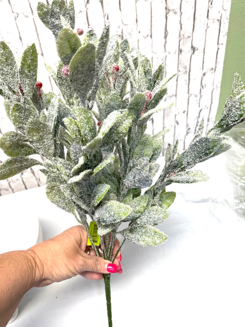 Puede incluir: Un ramo de vegetaci&oacute;n artificial con hojas verdes y peque&ntilde;as bayas rojas, cubierto con un material blanco similar a la nieve. Los tallos son verdes y las hojas son ovaladas. Fondo de pared de ladrillo blanco.