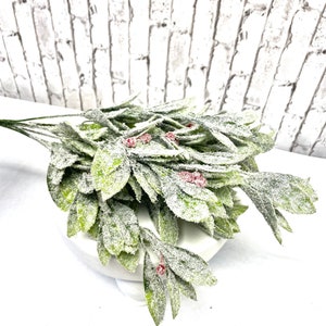 Puede incluir: Un ramo de vegetaci&oacute;n artificial con hojas verdes y peque&ntilde;as bayas rojas, cubierto con una sustancia blanca similar a la nieve. Las hojas est&aacute;n dispuestas en cascada, creando un aspecto natural y festivo. La vegetaci&oacute;n se coloca sobre un pedestal blanco.