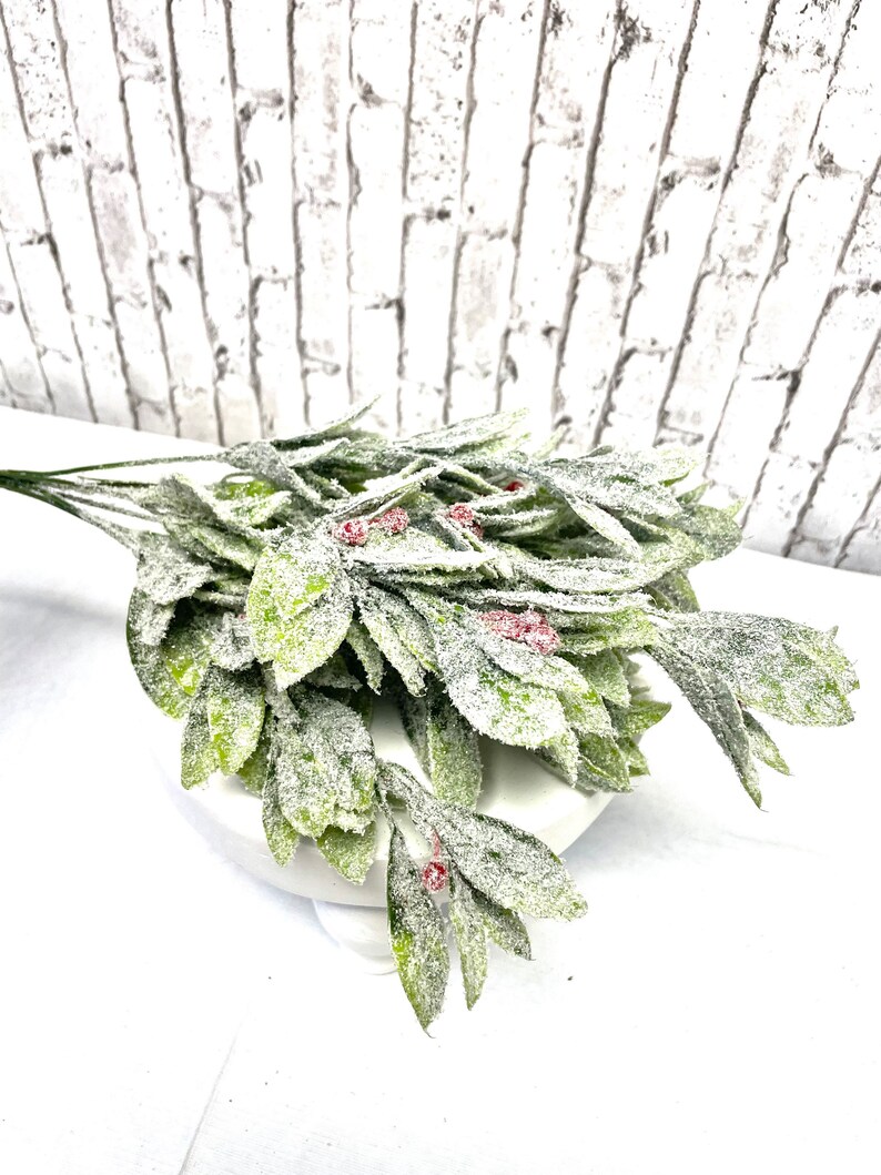 Puede incluir: Un ramo de vegetaci&oacute;n artificial con hojas verdes y peque&ntilde;as bayas rojas, cubierto con una sustancia blanca similar a la nieve. Las hojas est&aacute;n dispuestas en cascada, creando un aspecto natural y festivo. La vegetaci&oacute;n se coloca sobre un pedestal blanco.