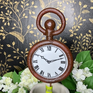 May include: A brown pocket watch with a white clock face and black Roman numerals. The watch has a loop at the top and is set against a floral background. The watch is approximately 5 inches in diameter.