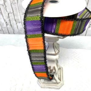 May include: Orange, purple, green, and black striped ribbon with black frayed edges. The ribbon is wrapped around a white spool.