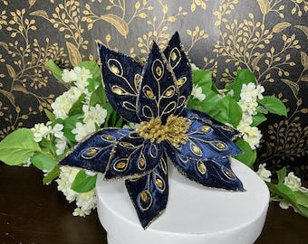May include: A large, navy blue velvet flower with gold accents and a gold center. The flower is decorated with gold leaf-like designs and gold-colored jewels. The flower is sitting on a white pedestal, with white flowers and green leaves in the background.
